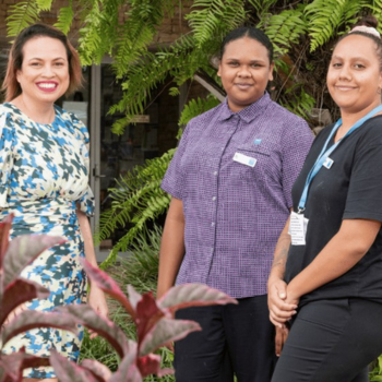 Three women stand together smiling outdoors among ferns and greenery, wearing name badges and lanyards.